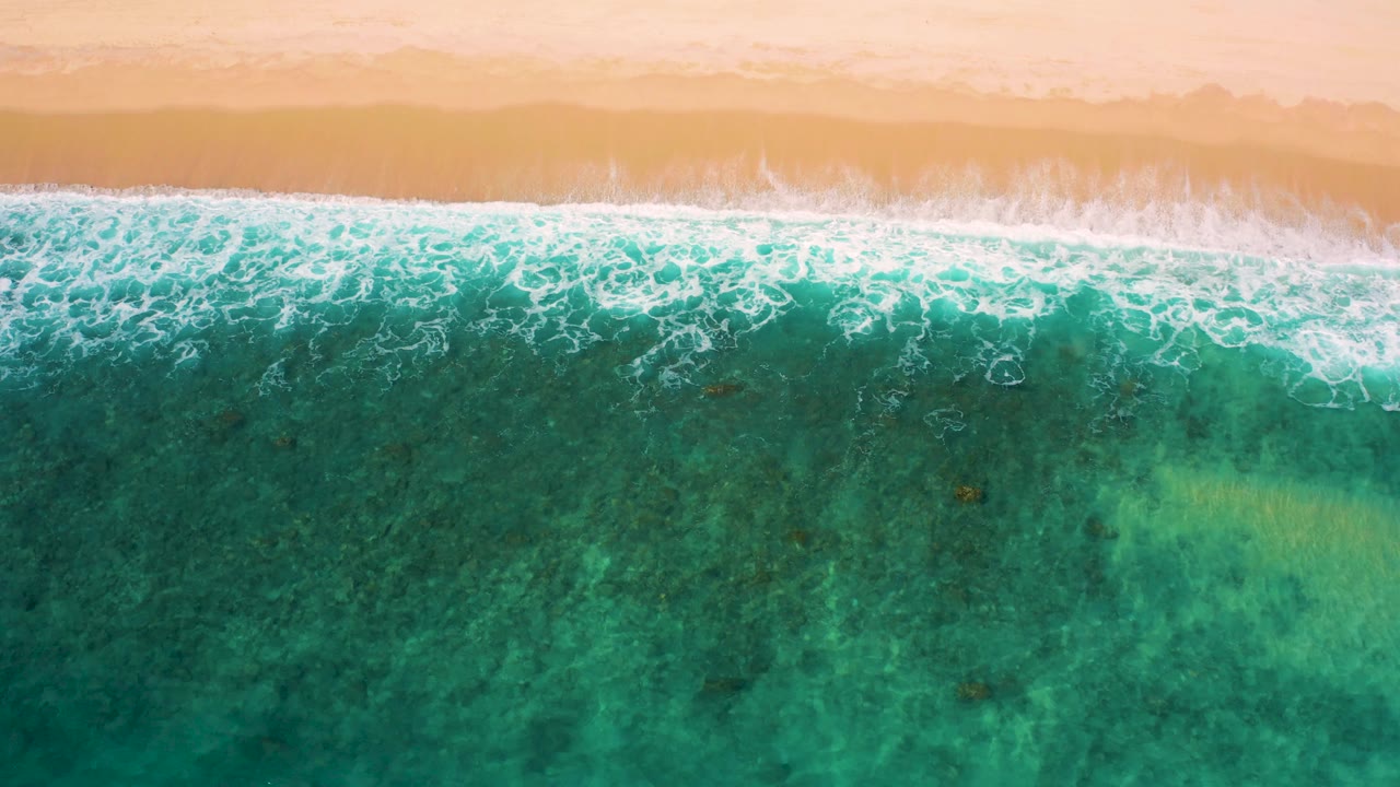 Top-down coastal view at El Cardoncito with a bright white surf band, peach sand above, clear teal water below, and no visible people.