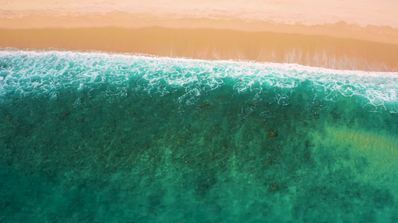 Aerial view of a diagonal foam line rolling along the sandy shore at El Cardoncito, with textured turquoise water and no visible people.