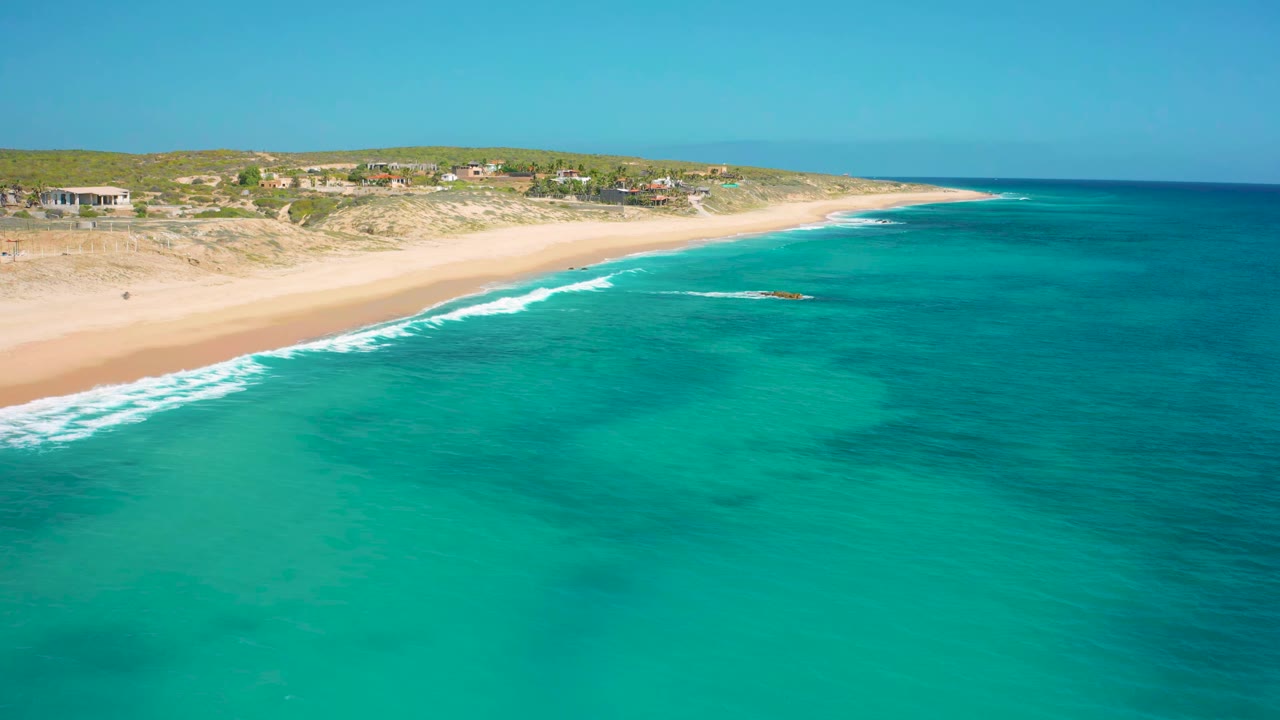 El Cardoncito shoreline seen from an aerial angle, with smooth turquoise water, a diagonal band of white surf, pale sand, low vegetation, and no visible people.