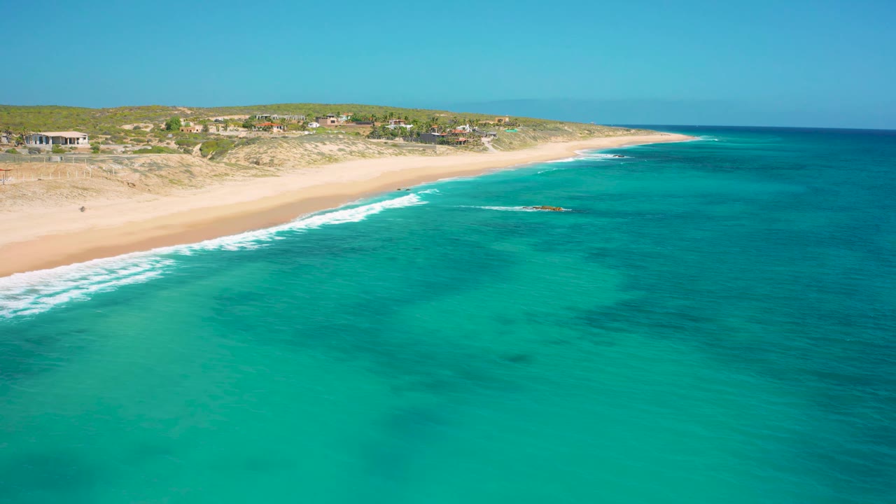 A wide aerial view of El Cardoncito with teal water across the foreground, a long pale beach, a few low coastal buildings beyond the dunes, and no visible people.