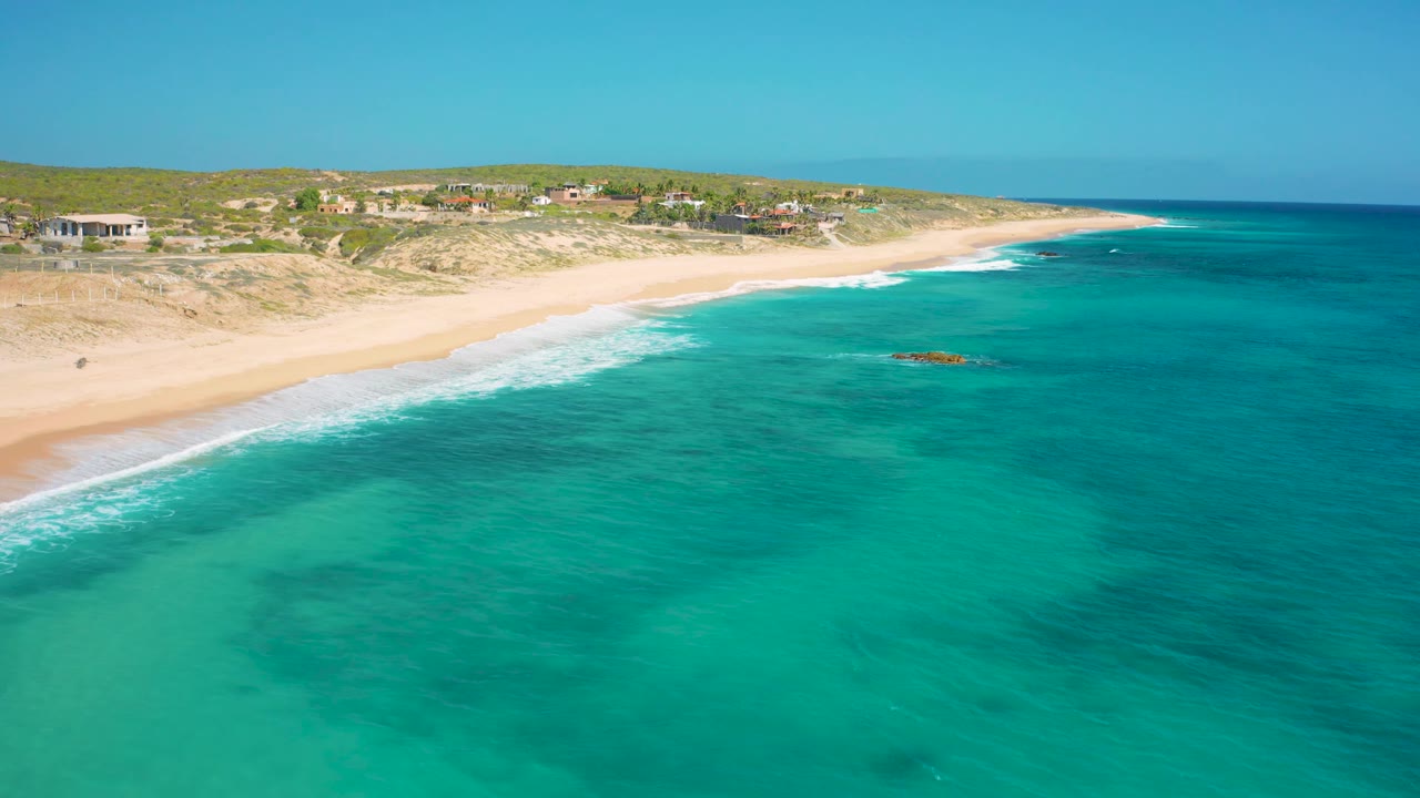 Aerial frame of the El Cardoncito coast with a small offshore rock visible in the turquoise water, white waves touching the empty beach, dunes behind the sand, and no visible people.