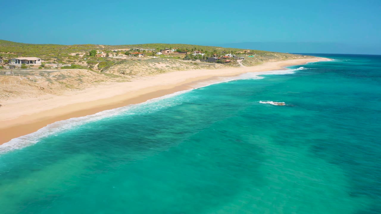 Aerial frame from El Cardoncito showing blue-green shallows, a long beach, white wave edges, dune vegetation, scattered coastal houses, and no visible people.