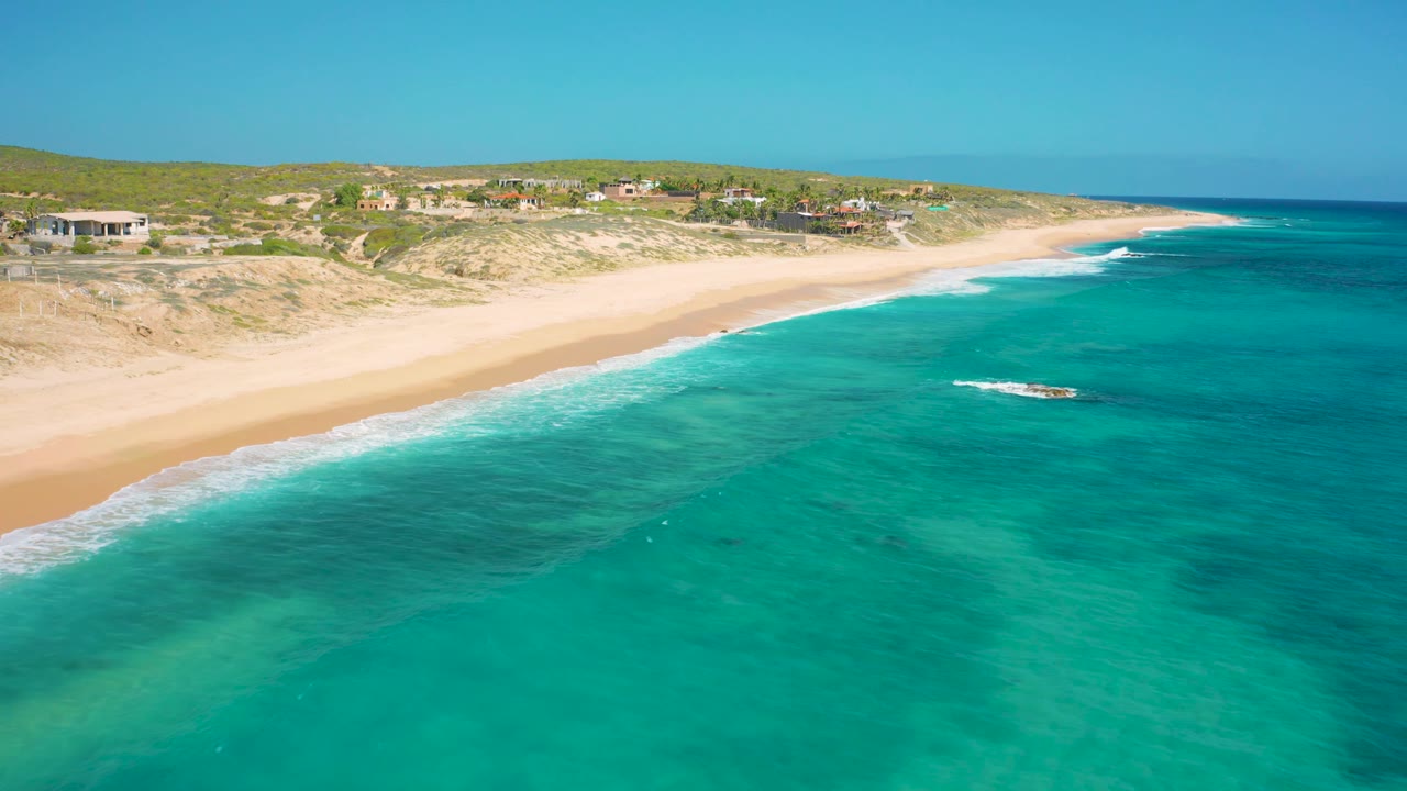Aerial view in El Cardoncito with turquoise water, a pale beach, a small rocky patch in the surf zone, low dunes, coastal homes, and no visible people.