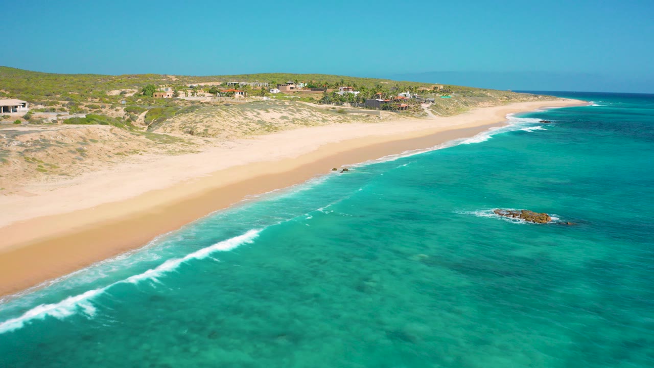 Aerial frame over El Cardoncito, Baja California Sur, showing clear turquoise water, a pale sandy shoreline, low dunes, scattered coastal buildings, and no visible people.