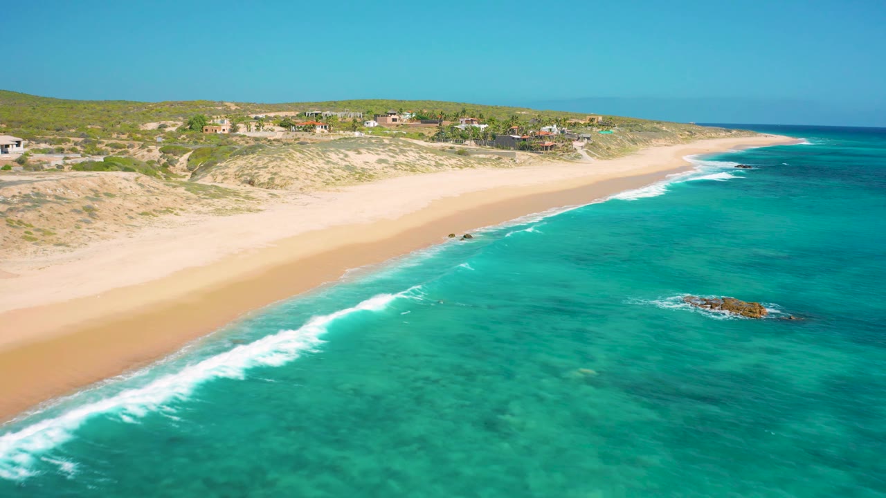 El Cardoncito shoreline seen from above with turquoise water filling the foreground, a small rocky outcrop offshore, pale beach, low green-brown dunes, distant buildings, and no visible people.