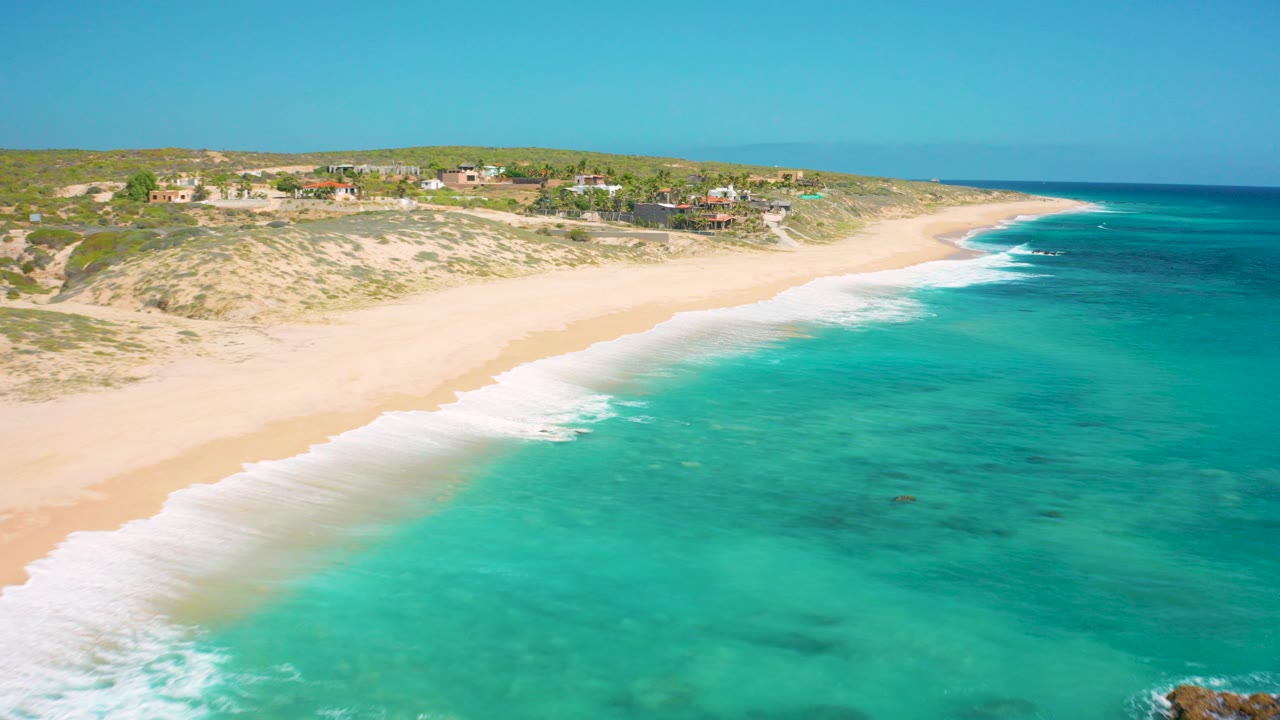 Aerial view over El Cardoncito, Baja California Sur, with turquoise water in the foreground, white surf on pale sand, low dunes, distant houses, and no visible people.