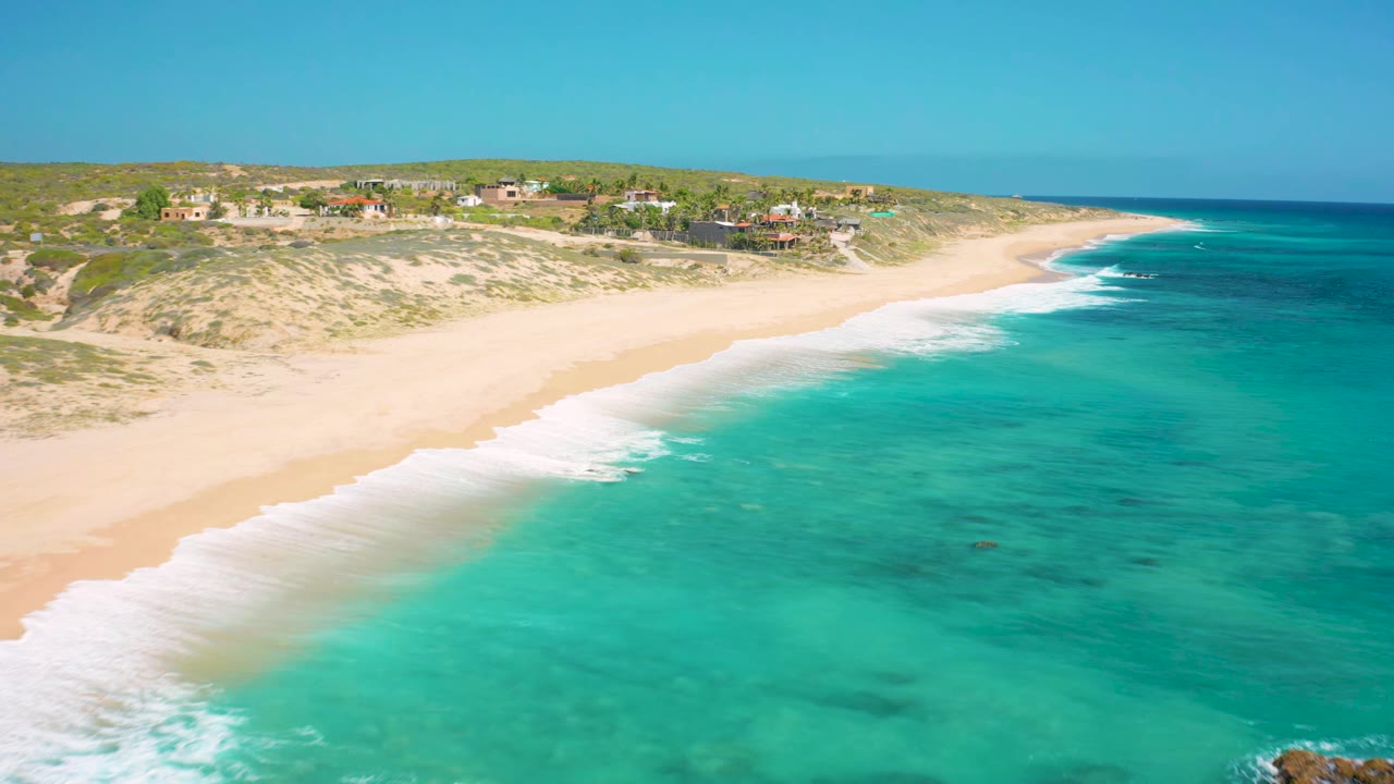 Second aerial frame over El Cardoncito shows a diagonal surf line, shallow turquoise water, pale sand, dune vegetation, distant coastal houses, and no visible people.