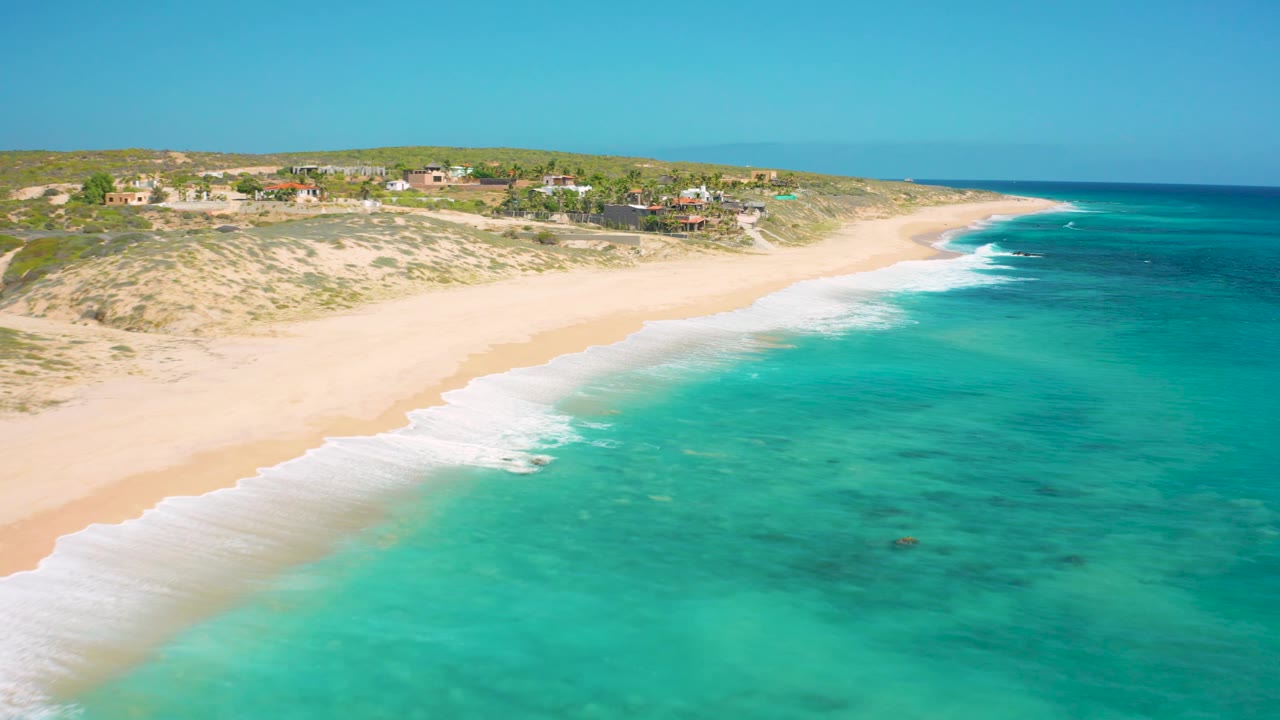 Aerial view of the El Cardoncito shoreline with more open water on the right, surf curling onto the sand, low dunes behind the beach, distant buildings, and no visible people.