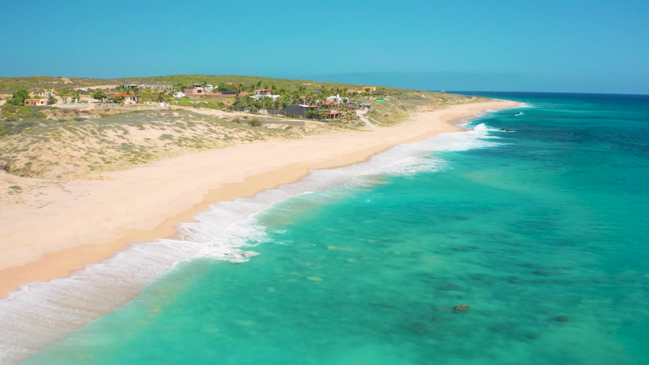 Wide aerial of empty sandy coast with turquoise shallows and surf washing up the beach.