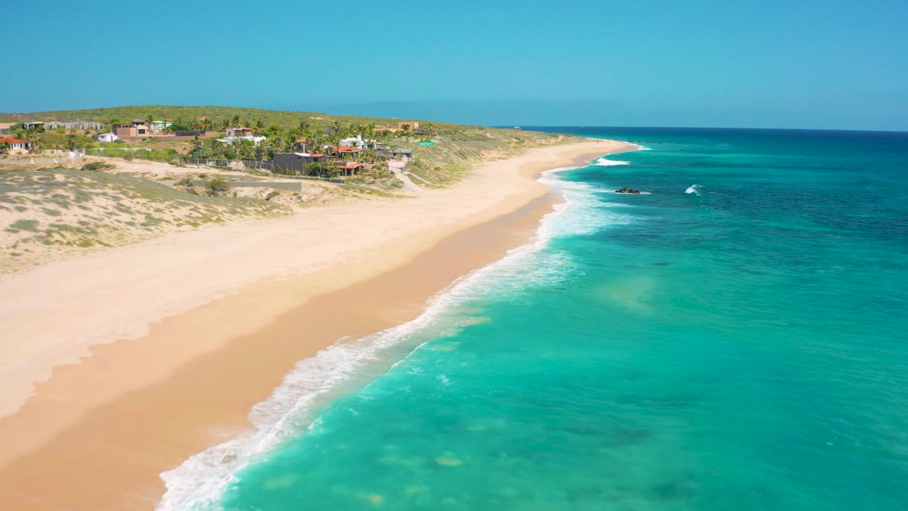Aerial view in El Cardoncito, Baja California Sur, with a wide sandy beach curving beside turquoise water and private homes on the bluff, no visible people.