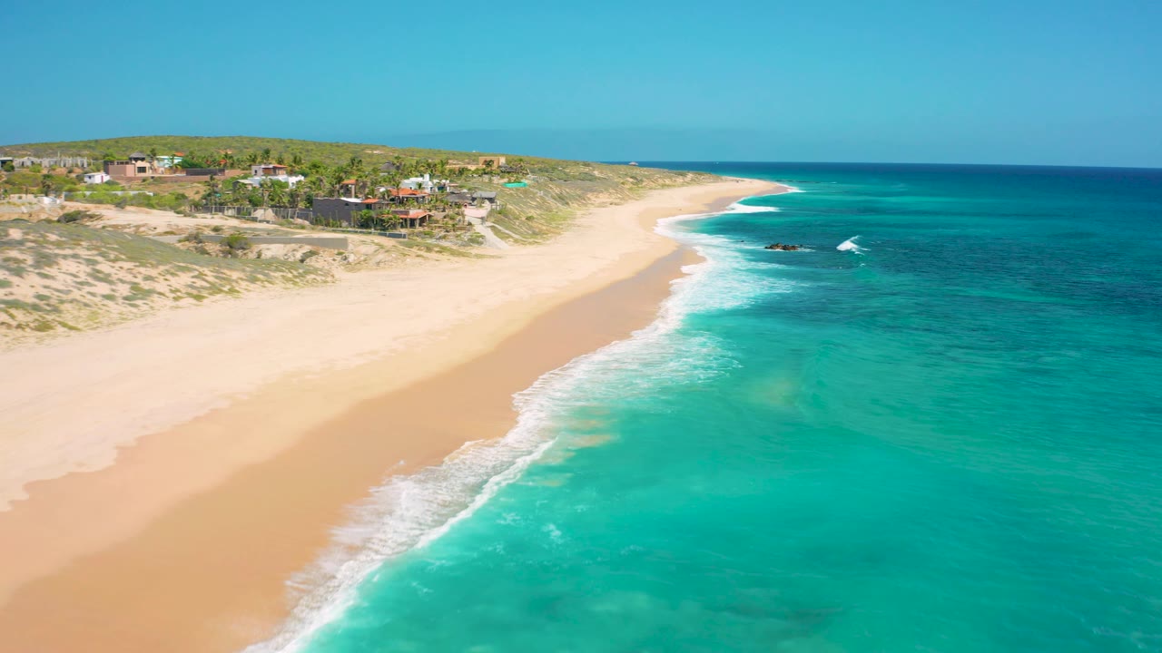 El Cardoncito beach fills the left side while turquoise water fills the right, with white foam near shore and private homes along the upper bluff, no visible people.