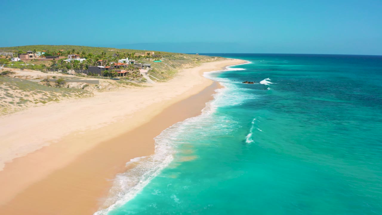 Coastal aerial with homes, sandy beach, turquoise sea, and a small breaking wave offshore.
