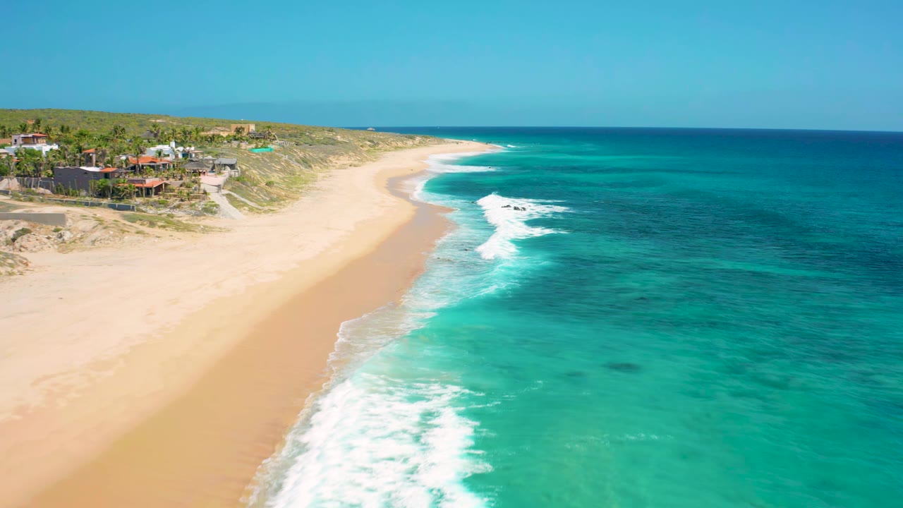 Wide aerial shoreline with clear blue water, white waves, beach, dunes, and houses.
