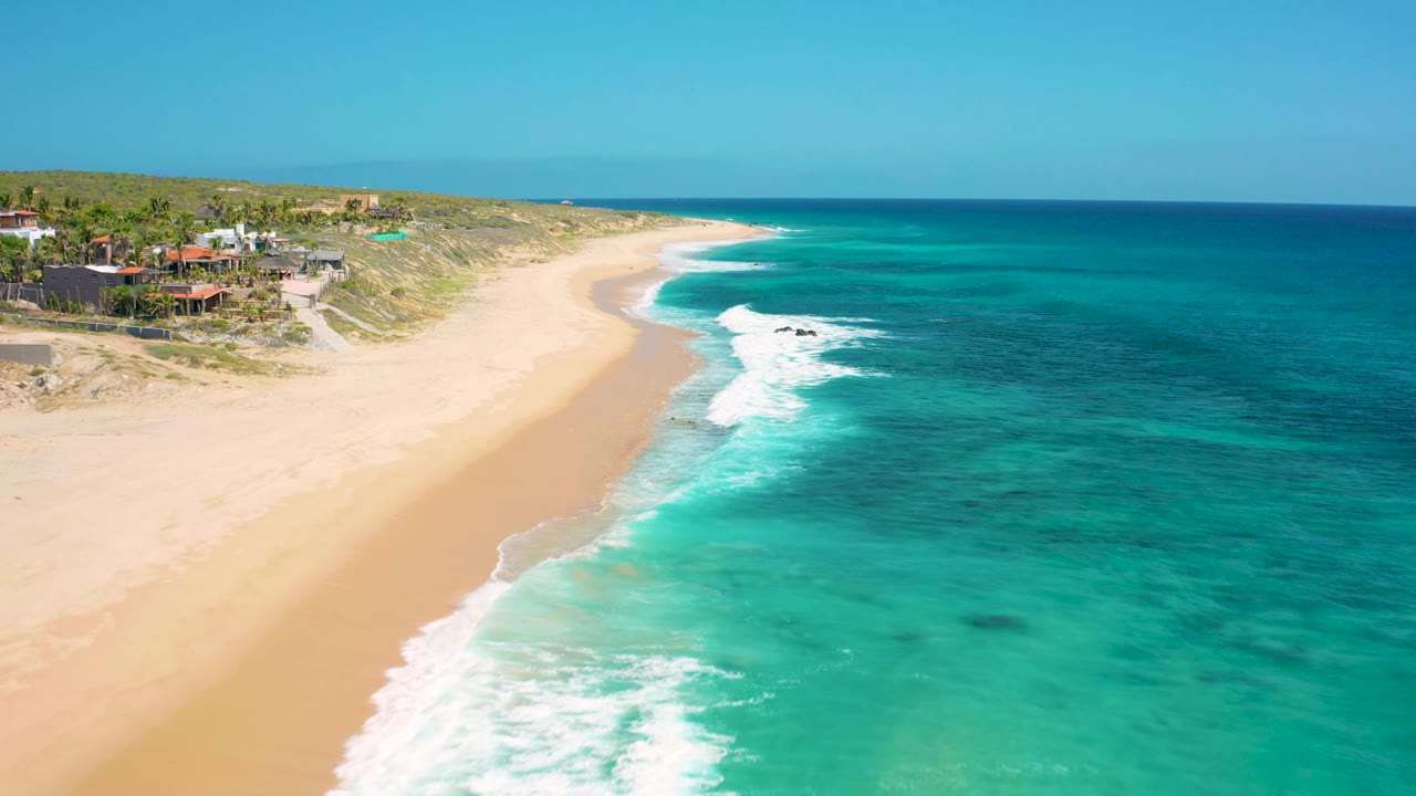 Aerial view along the El Cardoncito beach curve, showing brighter surf bands reaching the sand, shallow turquoise water near shore, darker blue water offshore, hillside vegetation, coastal structures, and no visible people.