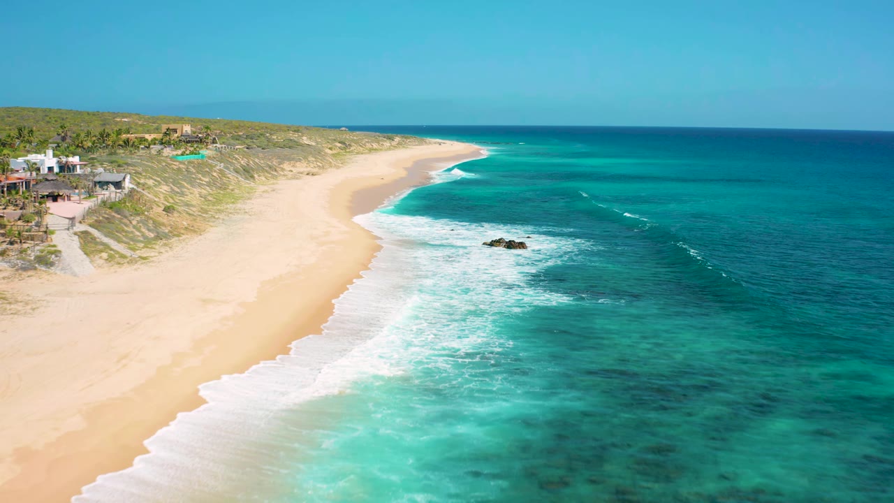 Aerial shoreline view with sandy beach, blue-green water, white waves, and dune homes.