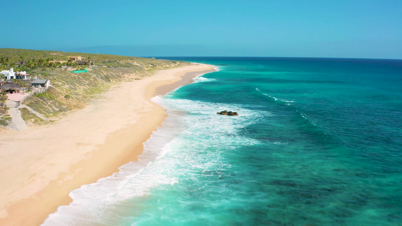 El Cardoncito aerial frame shows the curving sandbar-like beach edge, shallow turquoise water, darker offshore water, surf near rocks, dune homes, and no visible people.