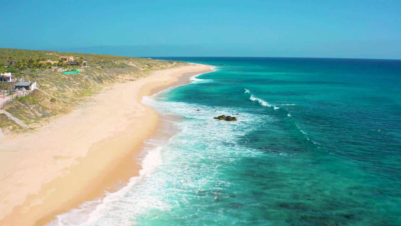 Aerial beach view with wide sand, foamy shoreline, rocks, dunes, and blue ocean.