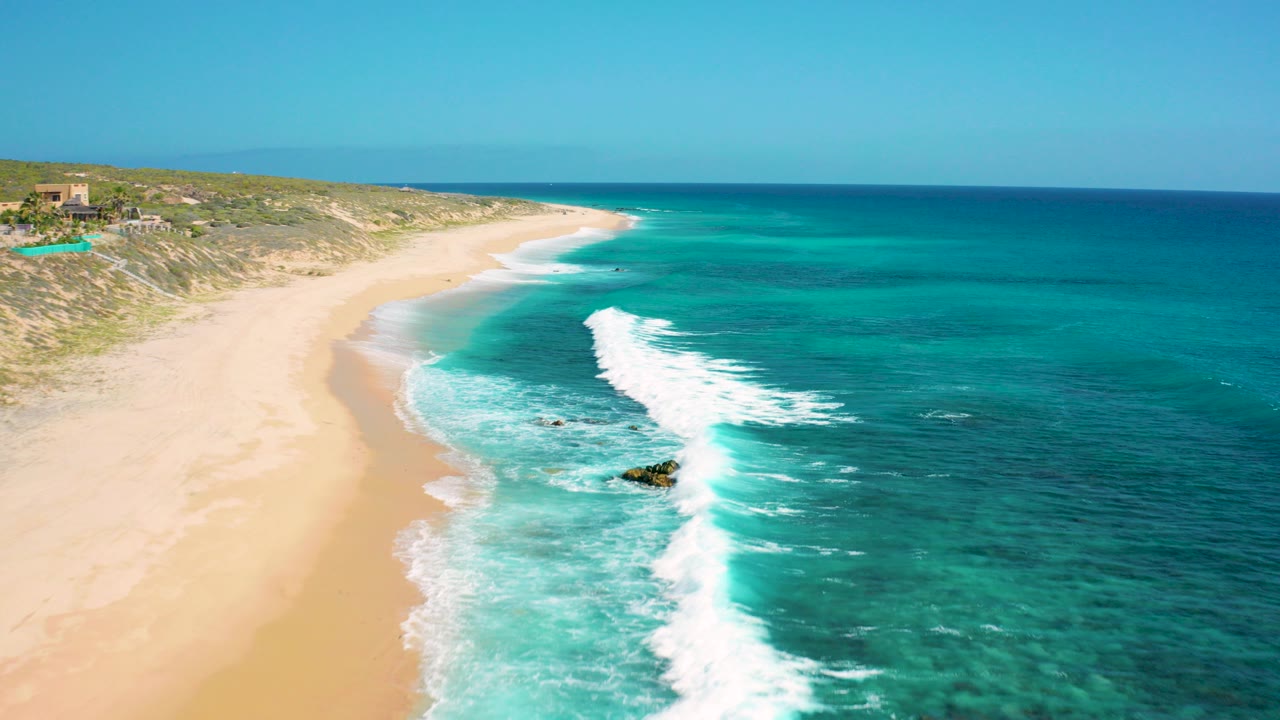 Aerial view over El Cardoncito, Baja California Sur, with turquoise waves breaking along a broad sandy beach, low dunes in the background, and no visible people.