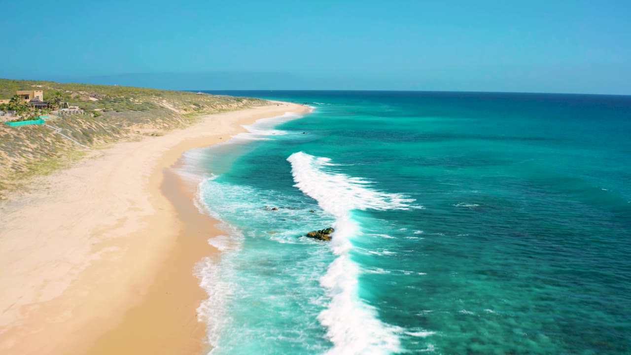 Aerial frame over El Cardoncito showing the beach curving into the distance while a white wave band crosses clear turquoise water, with no visible people.