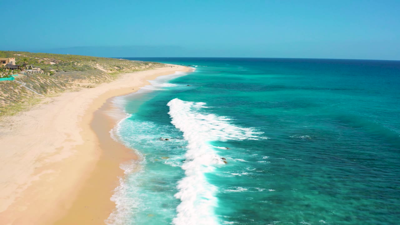 Aerial view at El Cardoncito with a foamy surf line running parallel to the empty tan beach and deeper blue water beyond, with no visible people.
