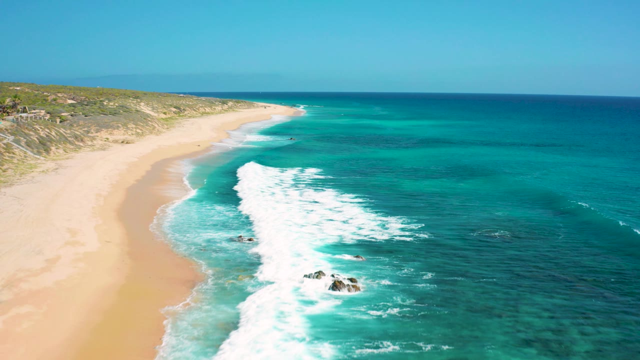 El Cardoncito shoreline from above, with bright foam breaking near small dark rocks in the shallow water and the empty beach stretching into the background, with no visible people.