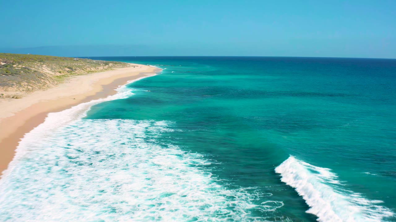 Aerial frame at El Cardoncito showing foamy waves pushing toward the pale sand while darker blue water sits beyond the reef-colored shallows, with no visible people.