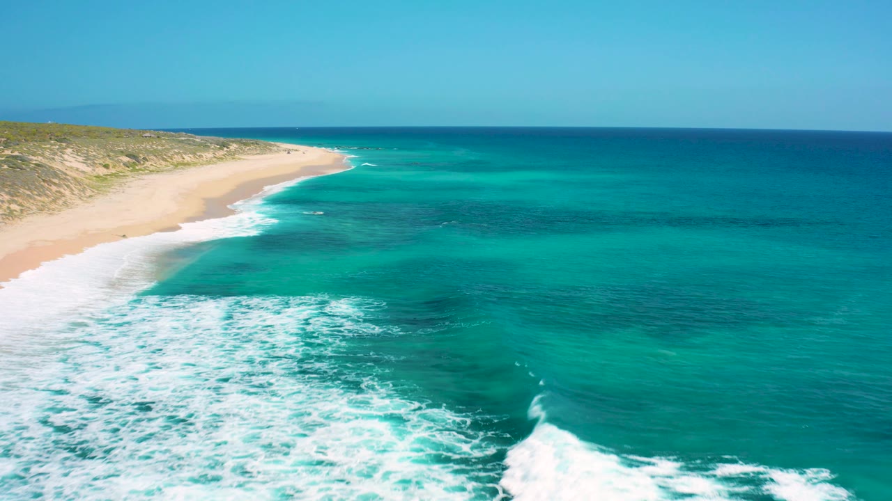 Aerial view over El Cardoncito with broad white surf foam in the lower frame, empty sand along the left edge, and open turquoise water extending to the horizon, with no visible people.