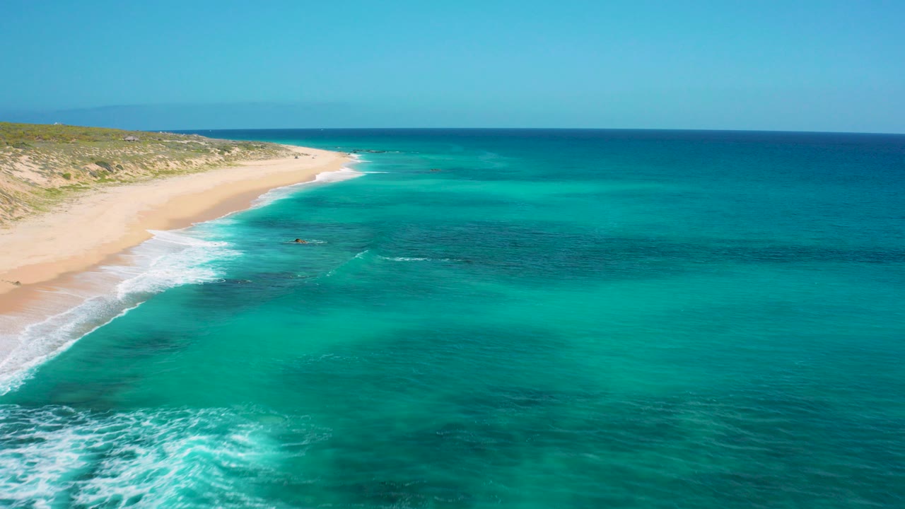 Aerial view over El Cardoncito showing turquoise water, white surf at the sand edge, low coastal vegetation behind the beach, and no visible people.