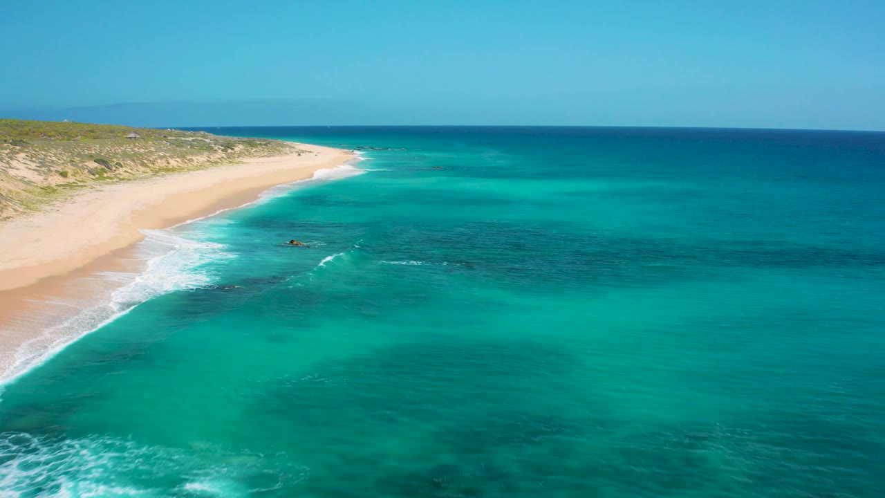 Aerial coastline at El Cardoncito with shallow turquoise water, darker reef-like patches offshore, a pale beach line at left, and no visible people.