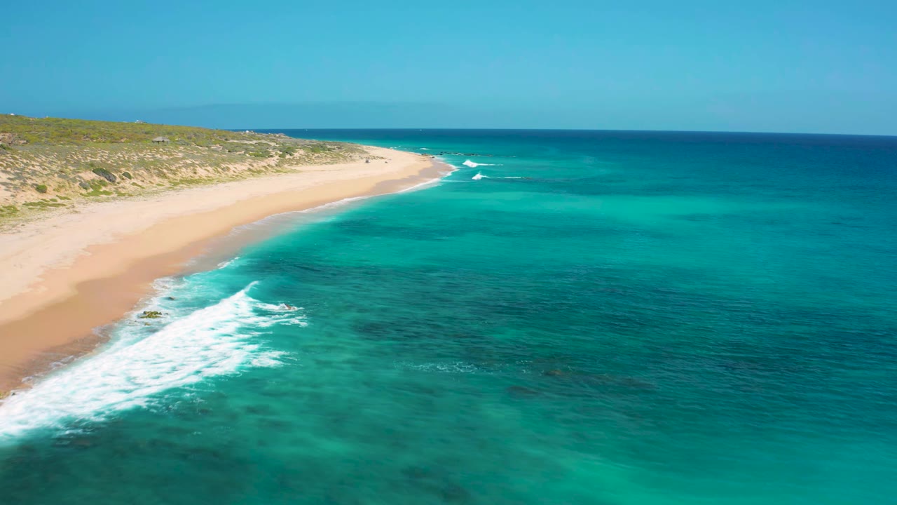 Aerial view at El Cardoncito showing a diagonal line of white surf washing toward a pale sand beach, with clear blue-green water offshore and no visible people.
