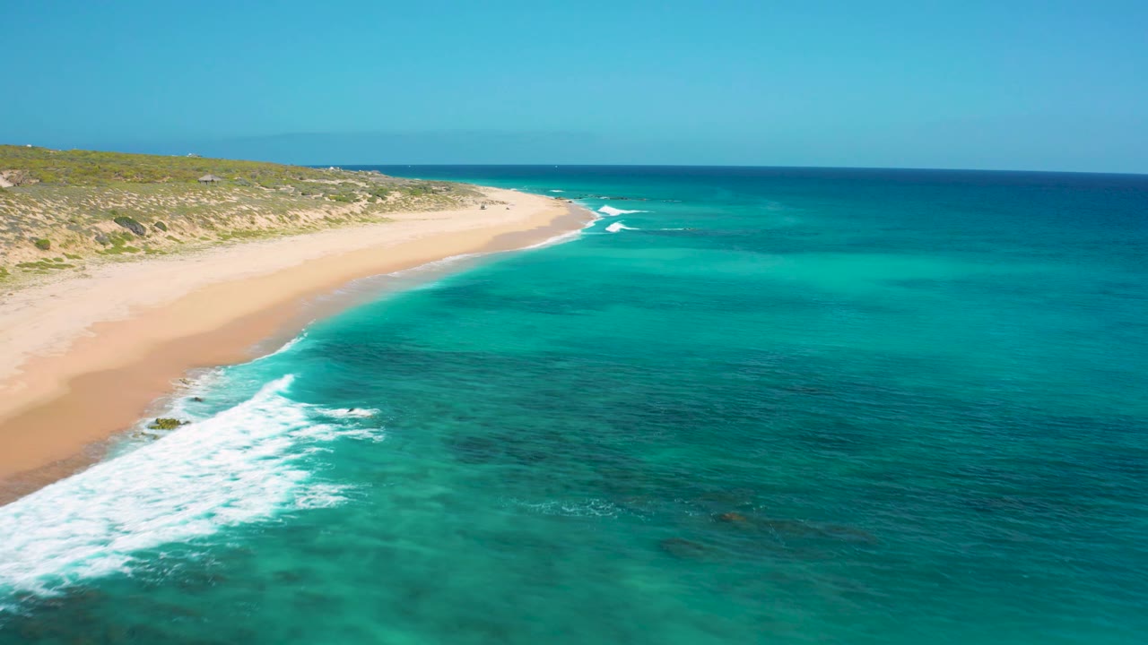 Drone frame over El Cardoncito with shallow turquoise water across the foreground, a curved sandy beach on the left, low coastal vegetation behind it, and no visible people.