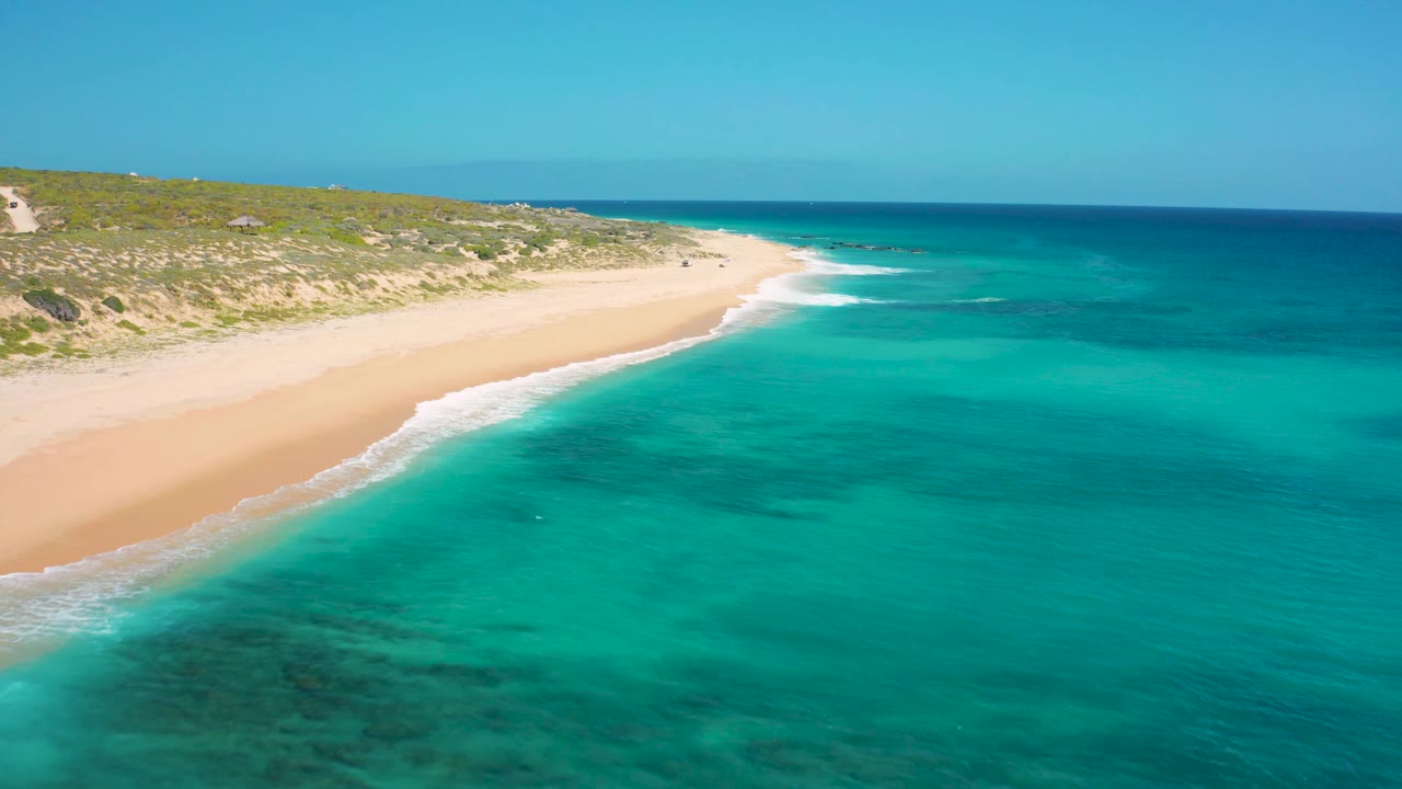 Drone-like frame over El Cardoncito showing blue-green ocean across the foreground, small white waves along the sand, low scrub-covered dunes, a clean horizon, and no visible people.