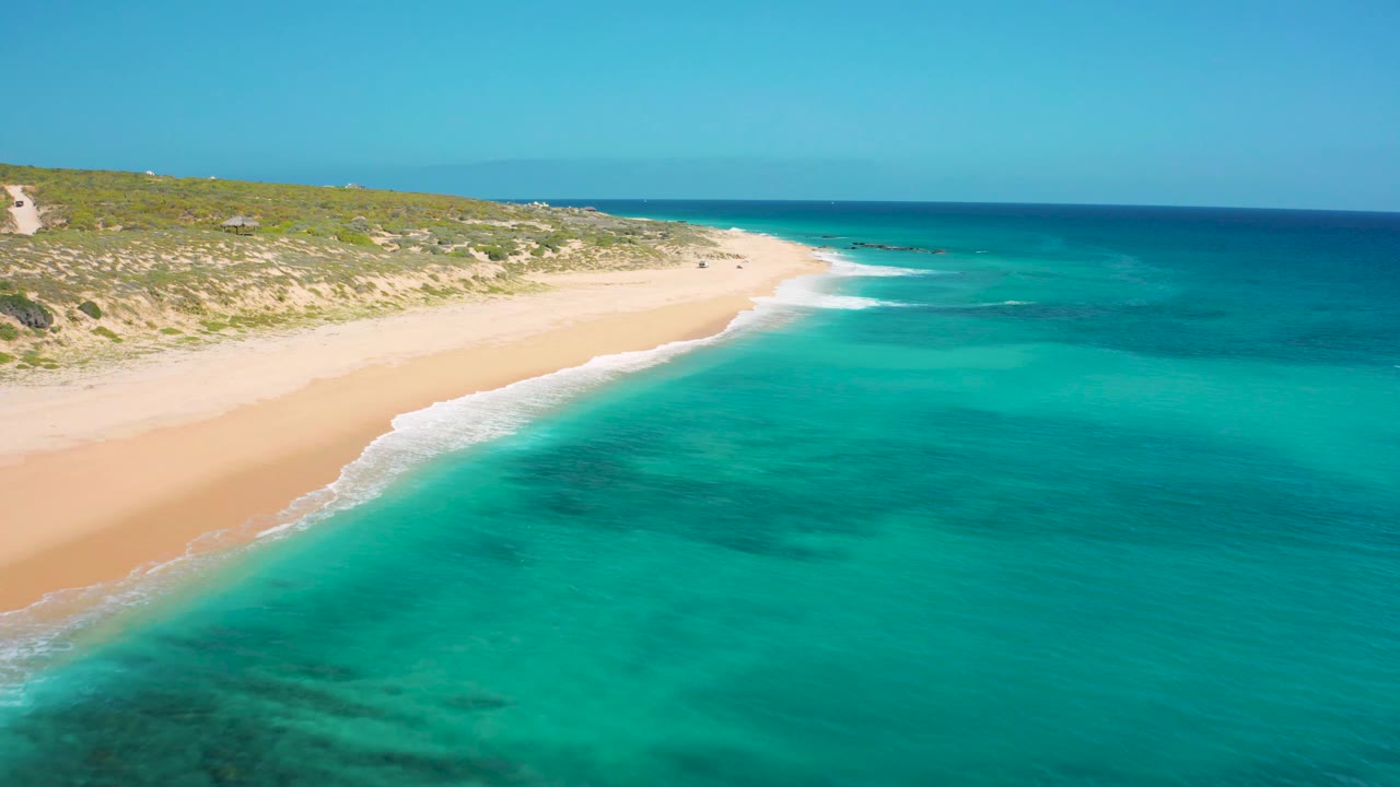 Wide aerial coastline with turquoise water, surf, sand, and dunes.