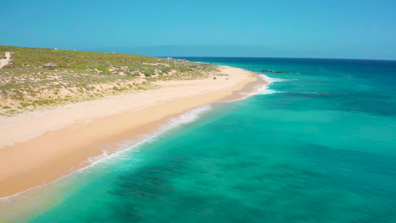 At 0:01, the El Cardoncito shoreline remains diagonal across frame with teal shallows in the foreground, deeper blue water beyond, dune scrub behind the sand, and no visible people.