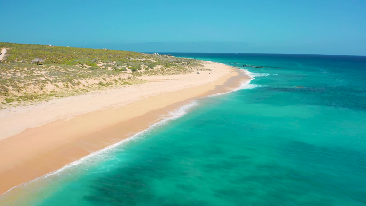 At 0:02, the aerial view shows the beach curve farther along the coast, with smooth sand, clear turquoise water, low vegetation inland, and no visible people.