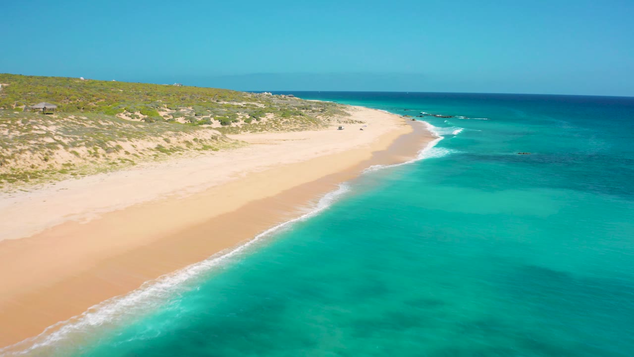 At 0:03, turquoise water fills the foreground while a pale sand strip and low dunes run across the upper left of the El Cardoncito frame, with no visible people.