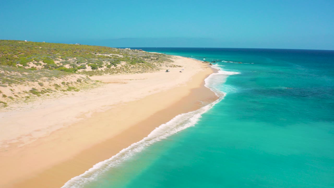 Aerial view of sandy beach beside turquoise water and green dunes.