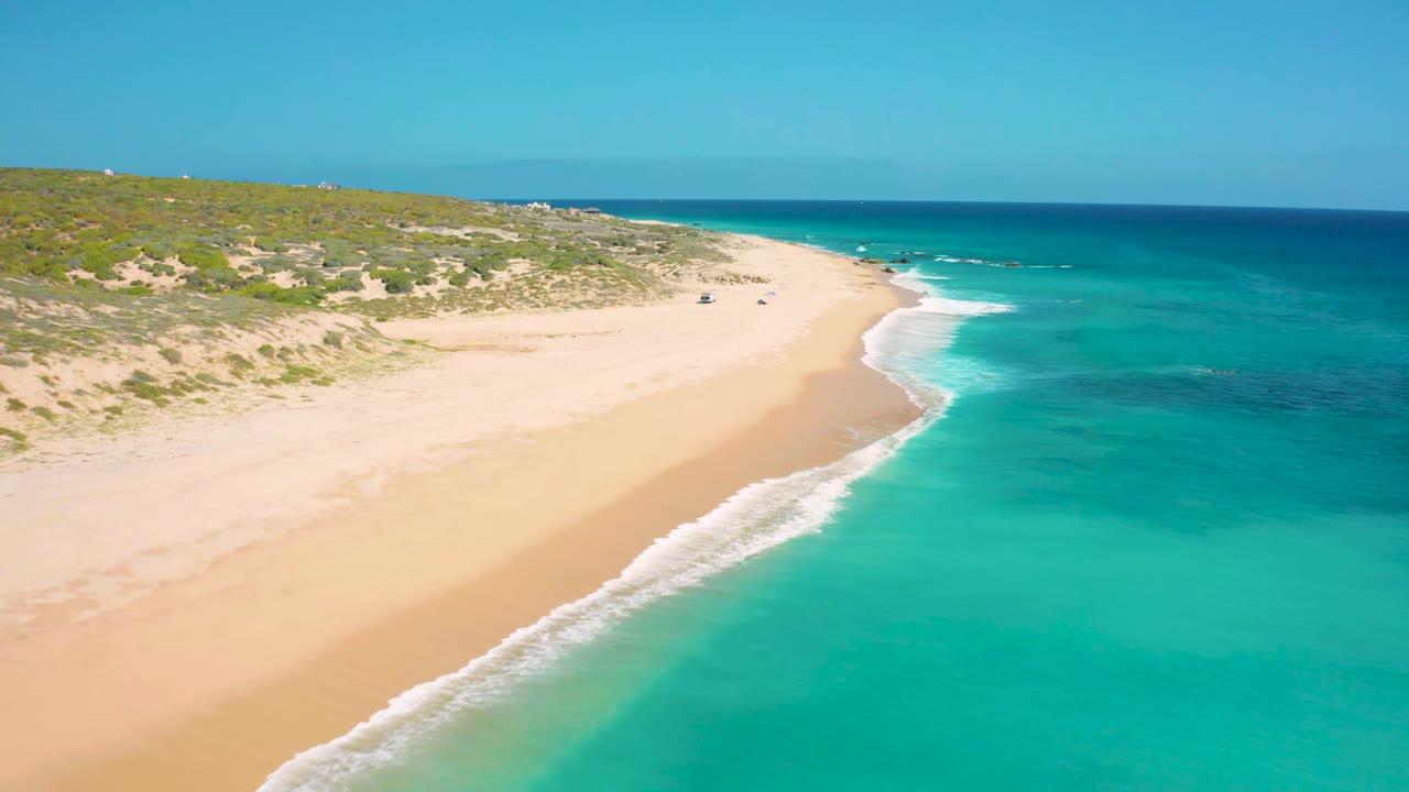 Aerial view along the El Cardoncito shoreline as white surf traces the sand, with turquoise water on the right, scrub vegetation inland, and no visible people.