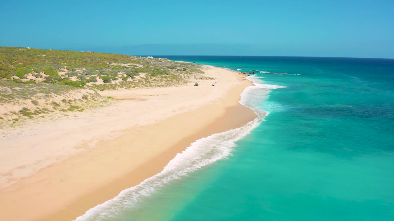 The beach curve at El Cardoncito fills the center of the frame, with clear blue-green water, a thin line of foam, low dunes behind the sand, and no visible people.
