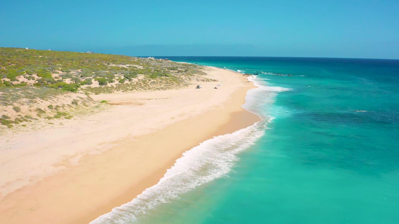 Mid-clip aerial frame over the El Cardoncito coast, showing the surf line lower in frame, wide empty sand, scrub-covered dunes, distant vehicles, and no visible people.