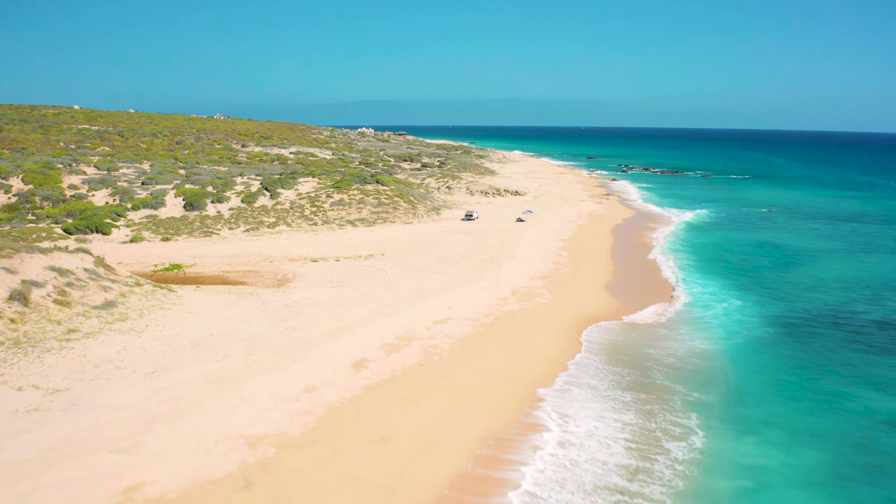 Aerial view at El Cardoncito with the shoreline running diagonally, low green dune vegetation on the left, bright surf on the right, and no visible people.