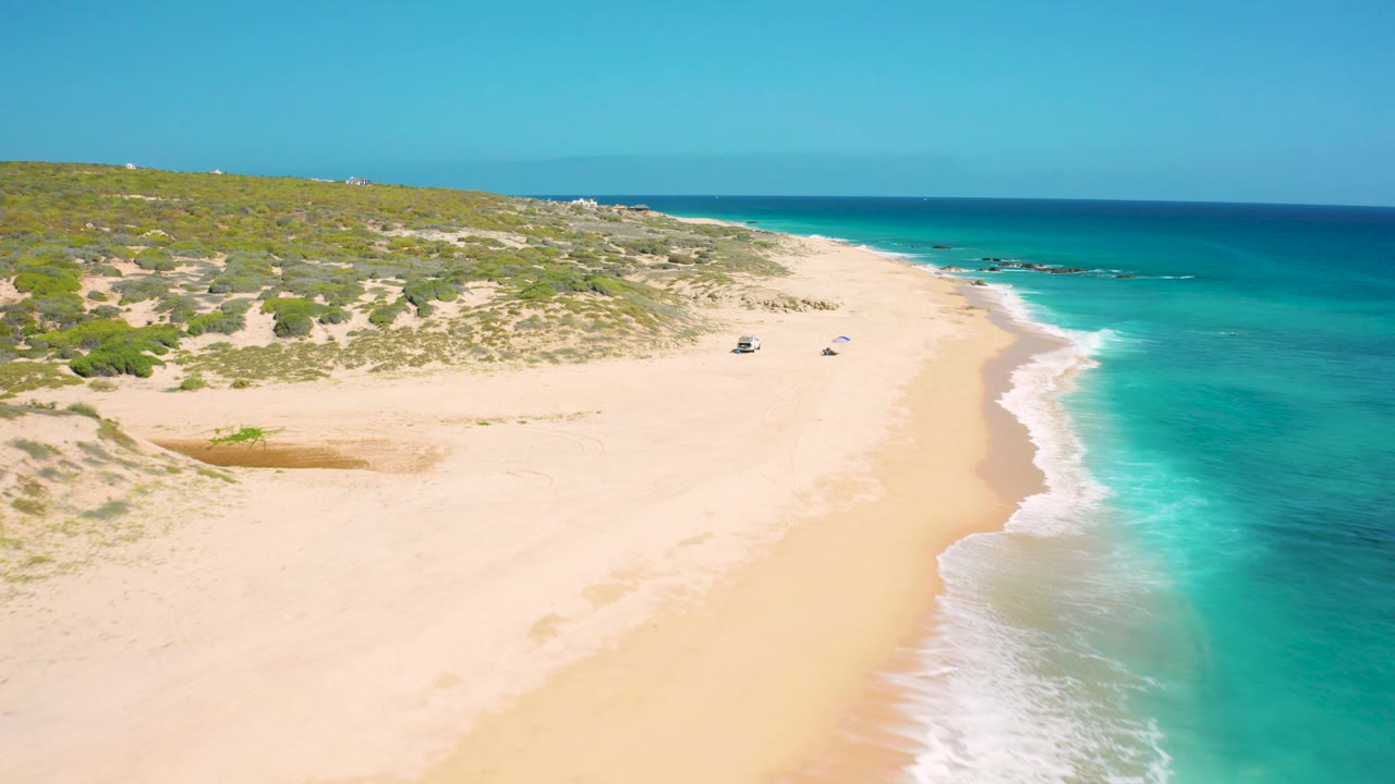 Aerial frame over the El Cardoncito beach with two small beach vehicles or objects on the sand, turquoise water close to shore, dunes in back, and no visible people.