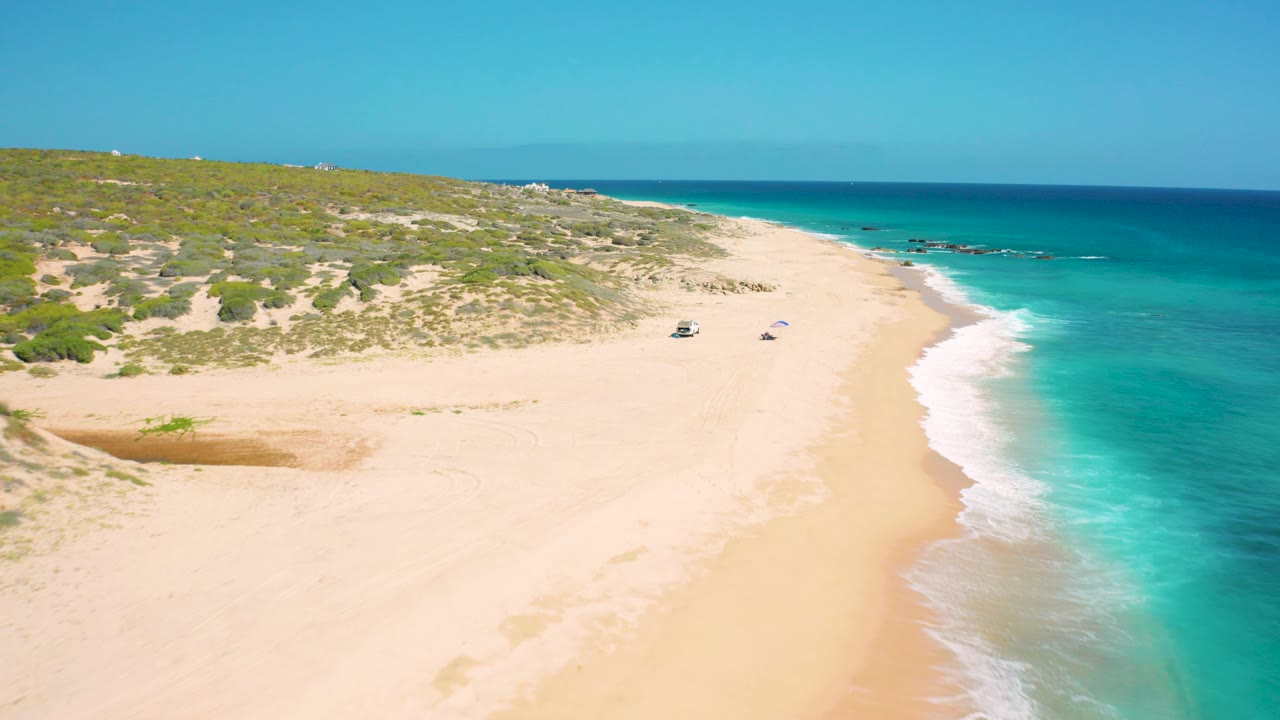Wide aerial view of El Cardoncito with a broad pale sand foreground, scrubby dunes behind it, a curved surf line on the right, and no visible people.
