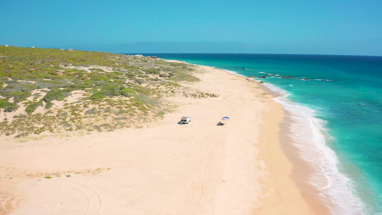 Aerial view of El Cardoncito beach with the surf line curving along the right side, scrub-covered dunes on the left, a vehicle and blue umbrella near the center sand, and no visible people.