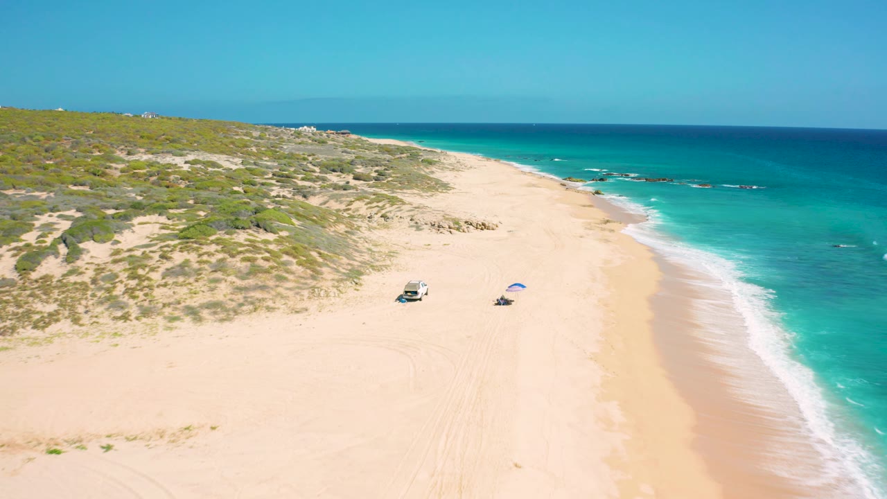 Aerial frame from El Cardoncito showing more of the foamy shoreline at right, pale beach below, dune vegetation to the left, a small vehicle and blue umbrella on the sand, and no visible people.