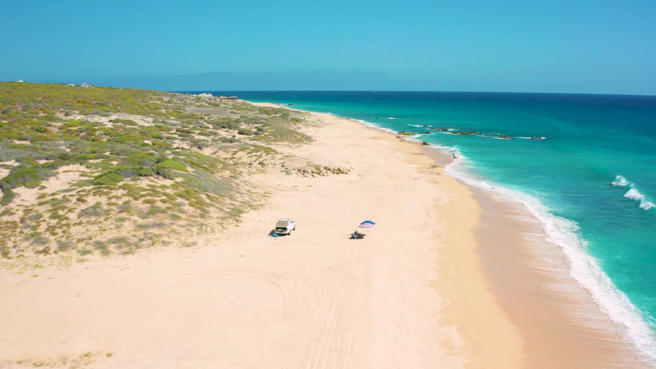 Mid-clip aerial view over El Cardoncito beach, with the vehicle and umbrella lower in frame, broad sand leading to low dunes, turquoise water along the right edge, and no visible people.