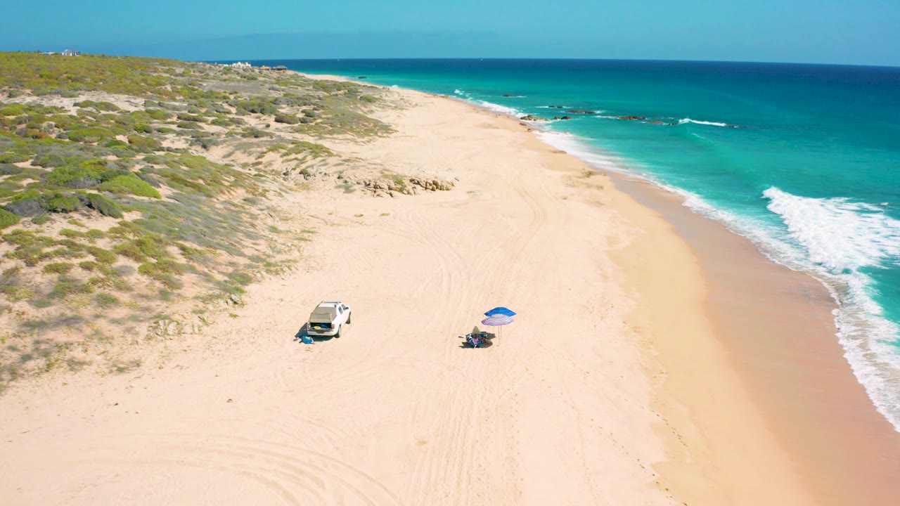 Aerial frame over El Cardoncito beach showing a white vehicle and blue umbrella set on pale sand, with low dunes to the left, turquoise water to the right, and no visible people.