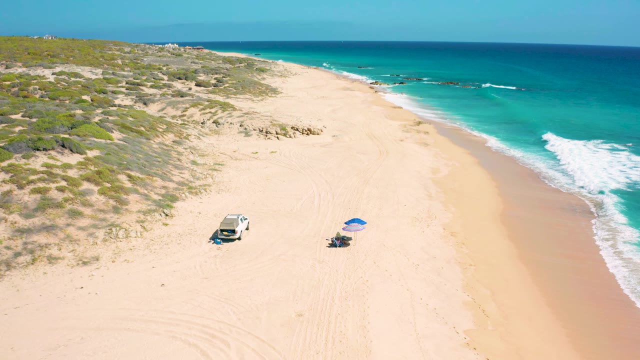 Elevated view at El Cardoncito with the parked white vehicle and blue umbrella slightly lower in frame, dunes filling the left side, ocean on the right, and no visible people.