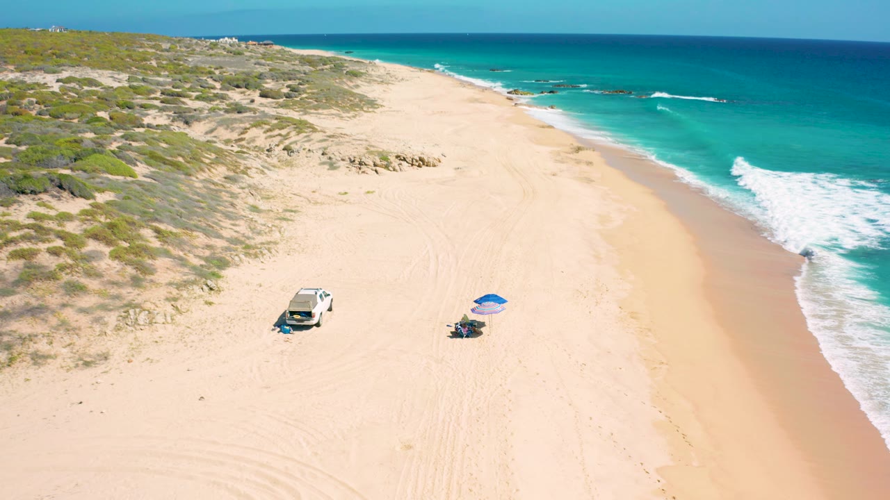 Wide aerial frame at El Cardoncito showing the beach curving into the distance, a white vehicle and blue umbrella on the sand, stronger surf at the right edge, and no visible people.