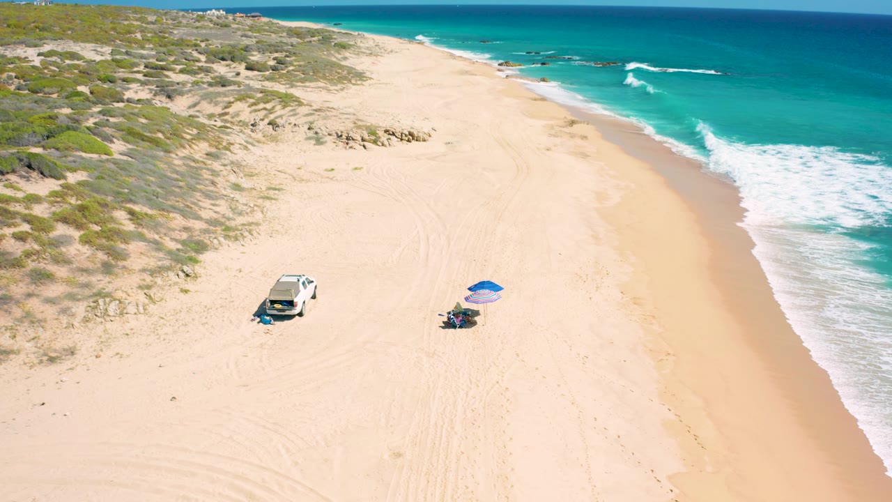 Drone view of empty sand with a vehicle, umbrella, dunes, and surf.